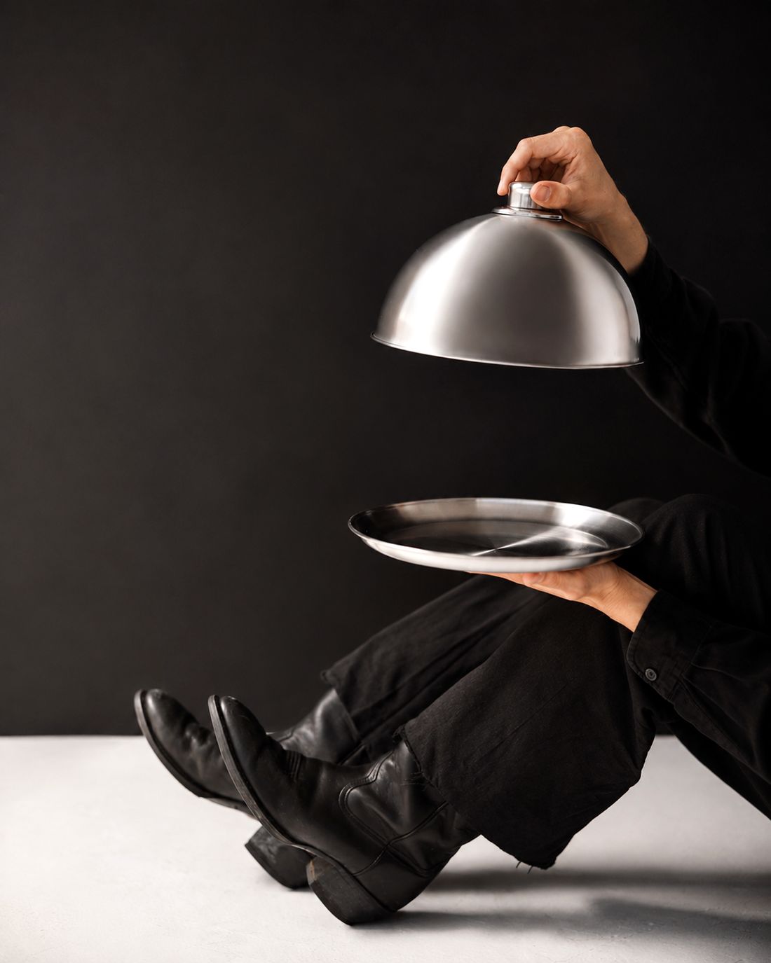 A server lifting a silver cloche from an empty platter against a black backdrop
